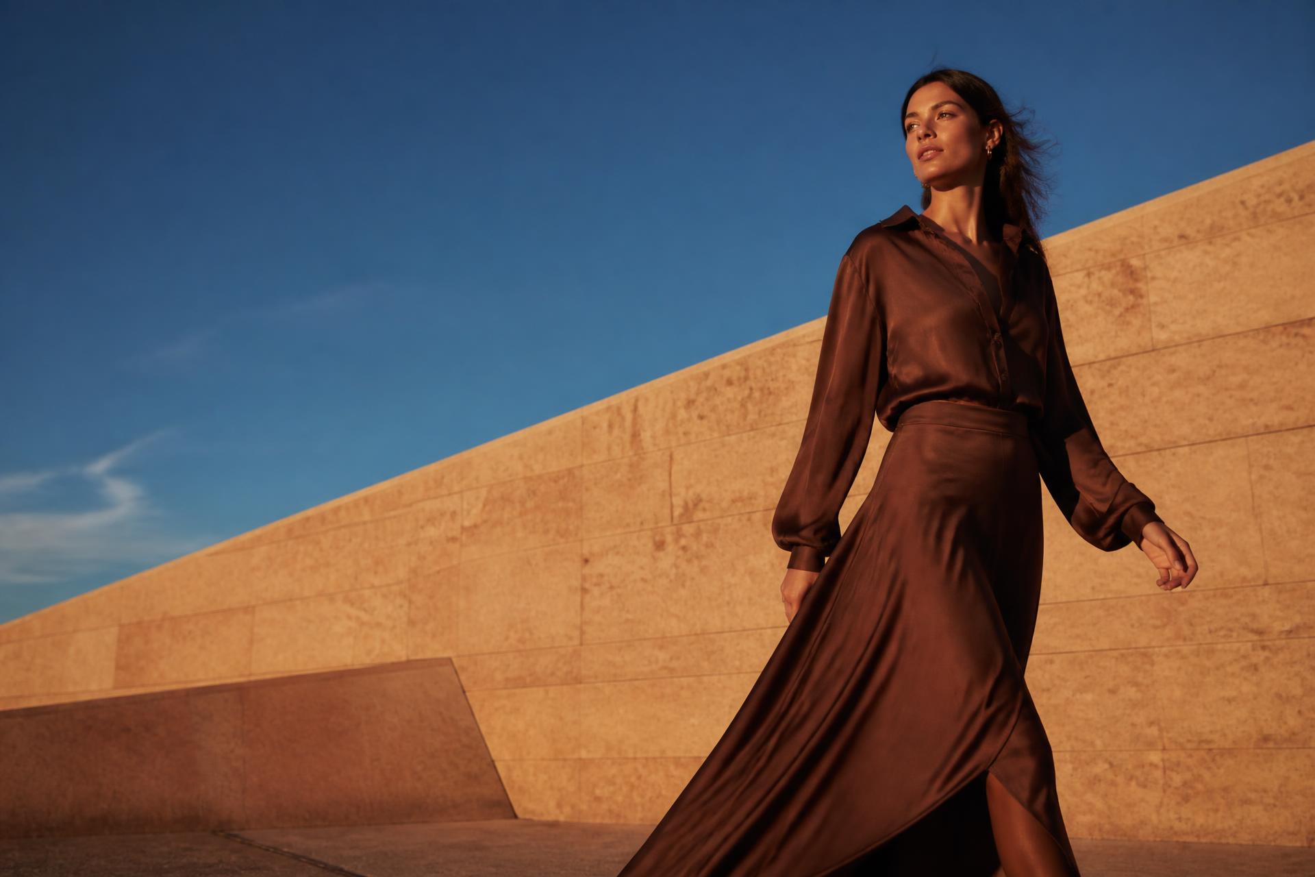 Model in a flowing brown silk shirt walking past a sunlit sandstone wall